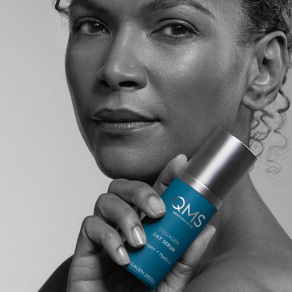 Eine Frau hält eine blaue Flasche QMS Collagen Day Serum nahe an ihr Gesicht und zeigt glatte, strahlende Haut auf einem Schwarz-Weiß-Foto mit dem Produkt in Farbe.