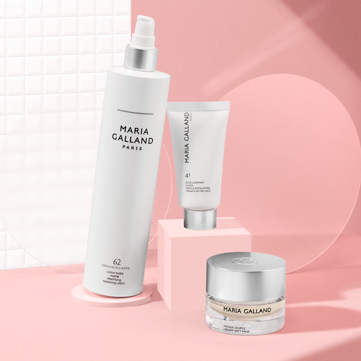 Drei Hautpflegeprodukte von Maria Galland Paris auf rosa-weißem Hintergrund: eine große Sprühflasche, eine Tube und ein kleiner Tiegel, stilvoll angeordnet mit minimalen Schatten.
