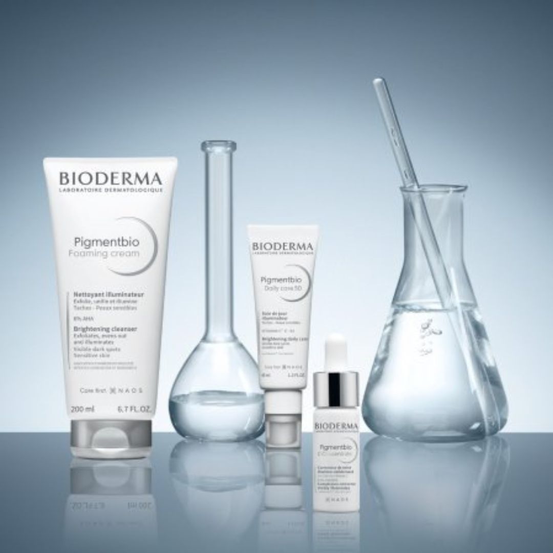 Die Hautpflegeprodukte von Bioderma Pigmentbio werden zusammen mit Laborgeräten aus Glas, darunter ein Becherglas, ein Kolben und eine Pipette, auf einer spiegelnden Oberfläche mit einem blauen Farbverlauf ausgestellt.