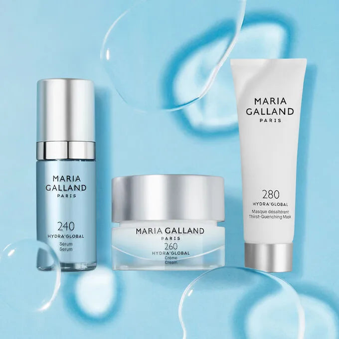 Drei Hautpflegeprodukte von Maria Galland Paris HydraGlobal - ein Serum, eine Creme und eine Maske - sind auf einem blauen Hintergrund mit Wassertropfen angeordnet, die Feuchtigkeit und Frische suggerieren.