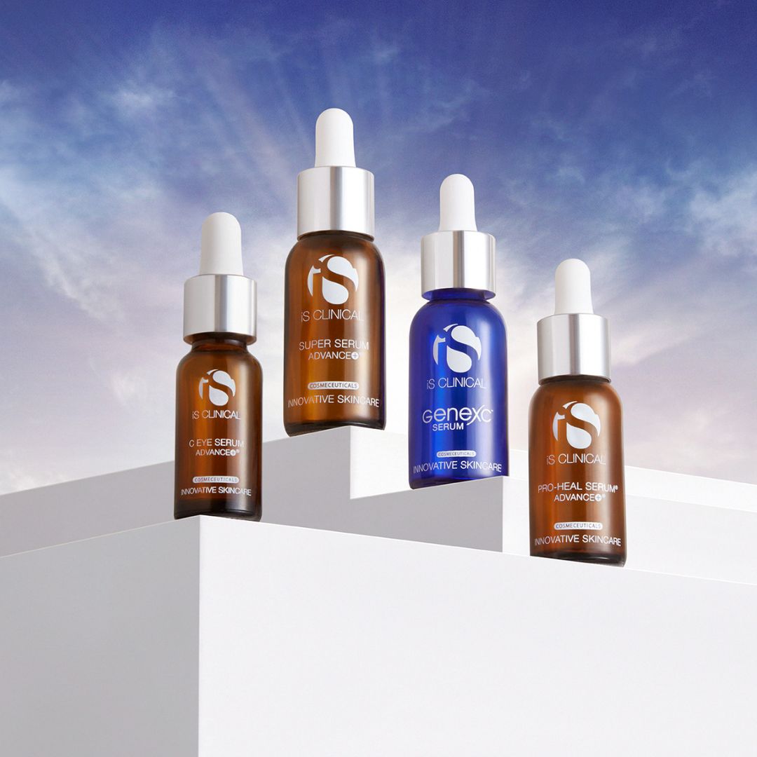 Vier Serumflaschen von iS CLINICAL, drei braune und eine blaue, sind auf weißen Podesten vor einem blauen Himmel mit Wolken ausgestellt. Jede Flasche ist mit einem Tropfverschluss und Markenetiketten versehen.