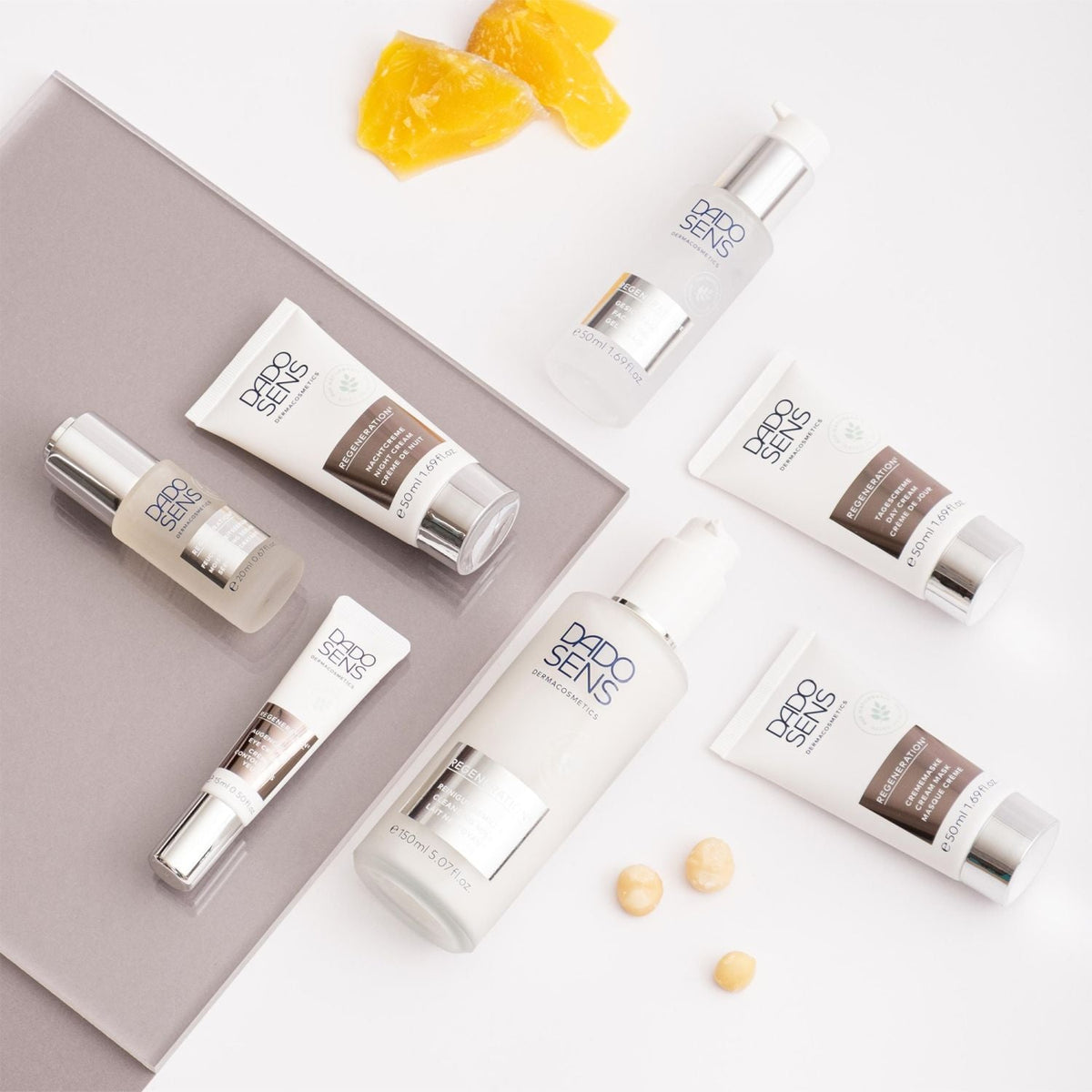 Eine flache Lage von DADO SENS Hautpflegeprodukten, einschließlich Lotionen und Cremes in weißen Flaschen und Tuben mit silbernen Kappen, angeordnet auf einem weißen und grauen Hintergrund mit gelbem Wachs und Macadamianüssen.