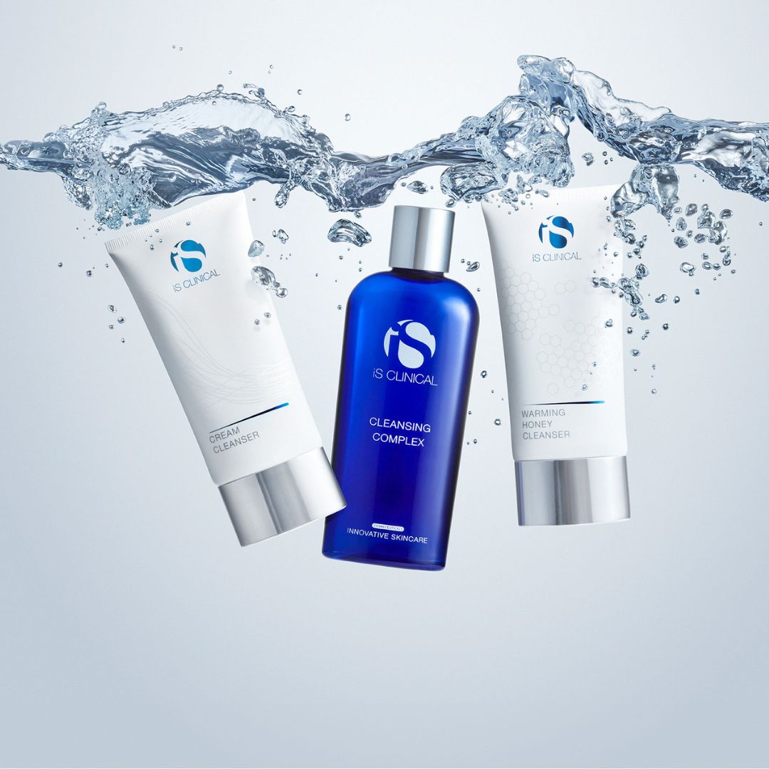 Drei Hautpflegeprodukte von iS Clinical - eine blaue Flasche Cleansing Complex und zwei weiße Tuben mit Reinigungsmitteln - sind vor einem hellen Hintergrund teilweise in spritzendes Wasser getaucht.