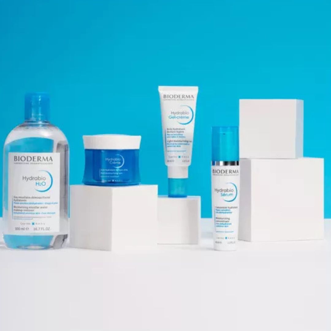 Vier Bioderma Hydrabio Hautpflegeprodukte sind auf weißen Blöcken vor einem blauen Hintergrund angeordnet, darunter ein Reinigungsmittel, eine Feuchtigkeitscreme, ein Serum und eine Tube Gel-Creme.