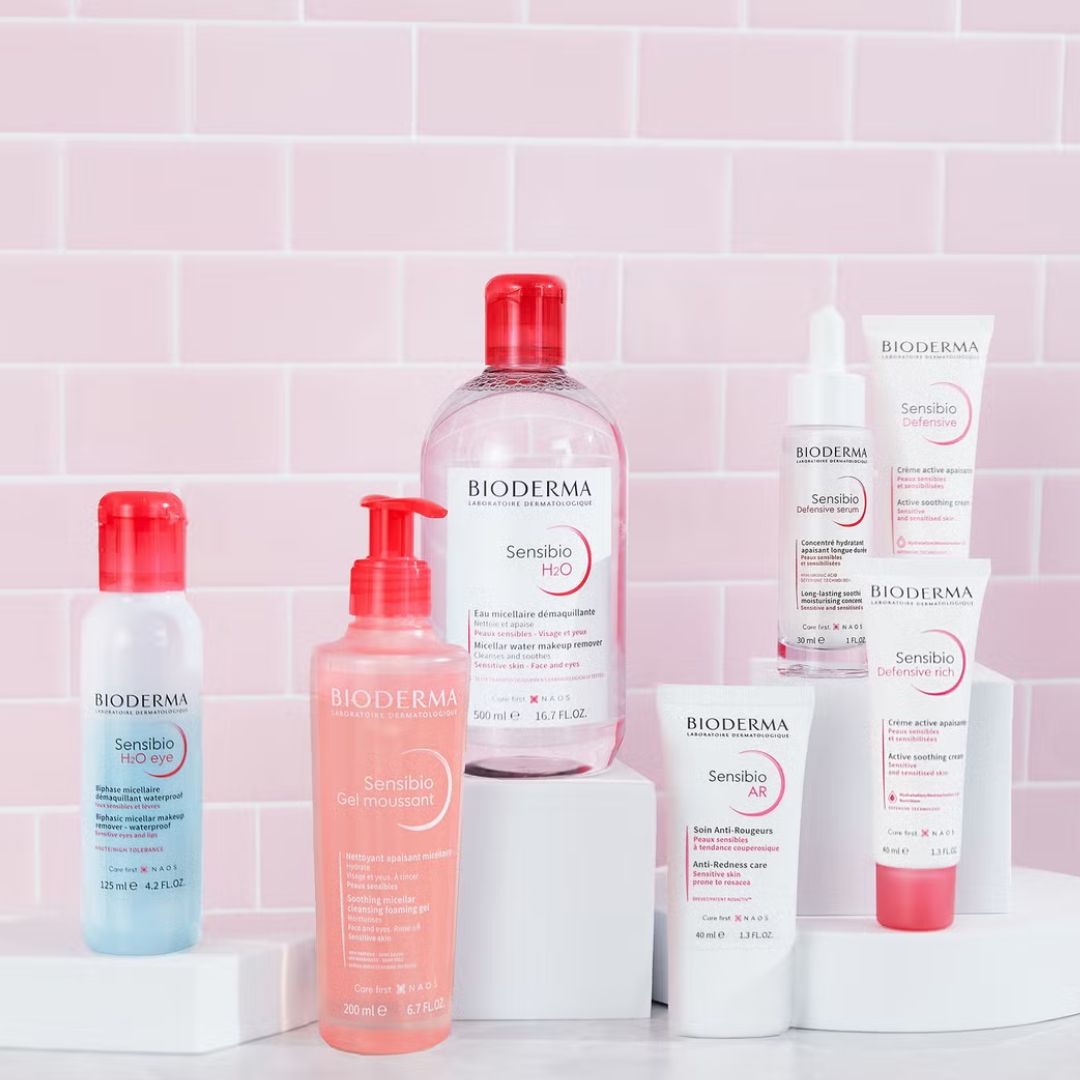 Sechs Hautpflegeprodukte von Bioderma, einschließlich Flaschen und Tuben, sind auf weißen Ständern vor einem rosa gekachelten Hintergrund ausgestellt. Die Produkte sind aus den Linien Sensibio und Sensibio AR.