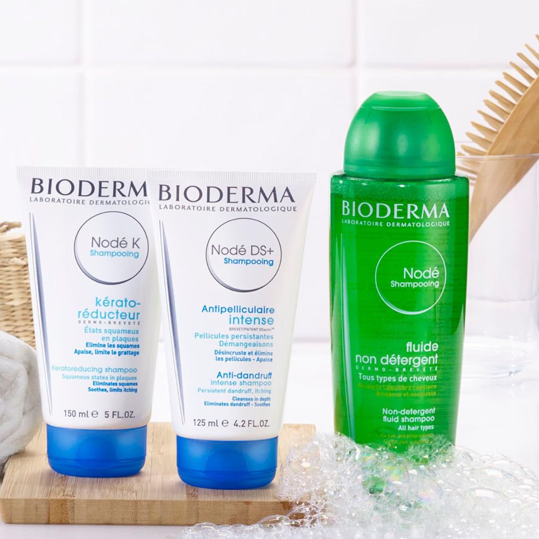 Drei Shampooflaschen von Bioderma stehen auf einer Theke mit Seifenlauge, einer Holzhaarbürste und einem Handtuch im Hintergrund. Die Flaschen sind mit Nodé K, Nodé DS+ und Nodé Shampooing beschriftet.