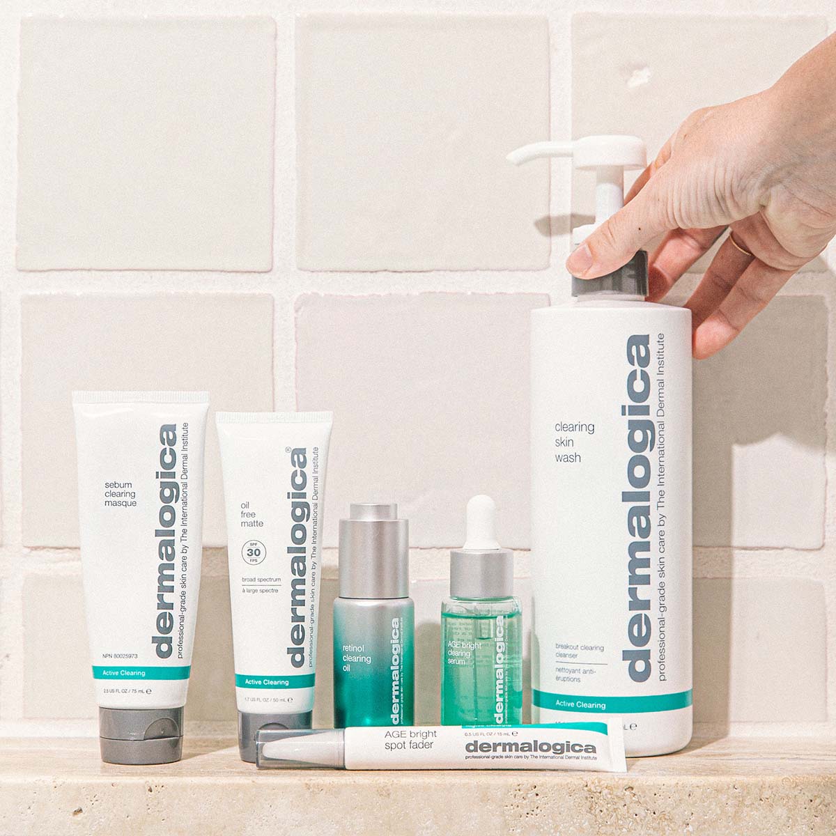 Eine Hand hält eine Pumpflasche mit Dermalogica Clearing Skin Wash, umgeben von verschiedenen Dermalogica-Hautpflegeprodukten, die auf einem gekachelten Badezimmerregal angeordnet sind.