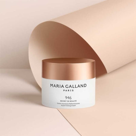 Ein Cremetiegel Maria Galland Paris 946 Secret de Beauté mit roségoldenem Deckel steht auf einer beigen Fläche mit passendem geschwungenem Hintergrund.