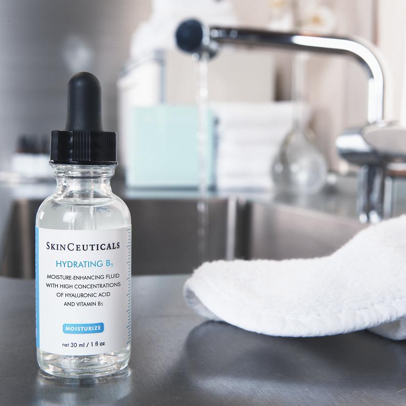 Eine Flasche SkinCeuticals Hydrating B5 Serum mit einer Pipette steht auf einem Badezimmertisch neben einem weißen Handtuch, während im unscharfen Hintergrund ein Wasserhahn läuft.