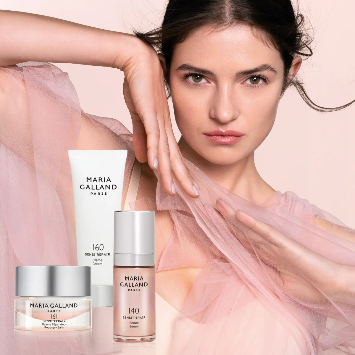 Eine Frau in einem durchsichtigen rosafarbenen Oberteil posiert mit erhobenem Arm neben drei Hautpflegeprodukten von Maria Galland Paris - Creme, Lotion und Serum -, die im Vordergrund vor einem zartrosa Hintergrund abgebildet sind.