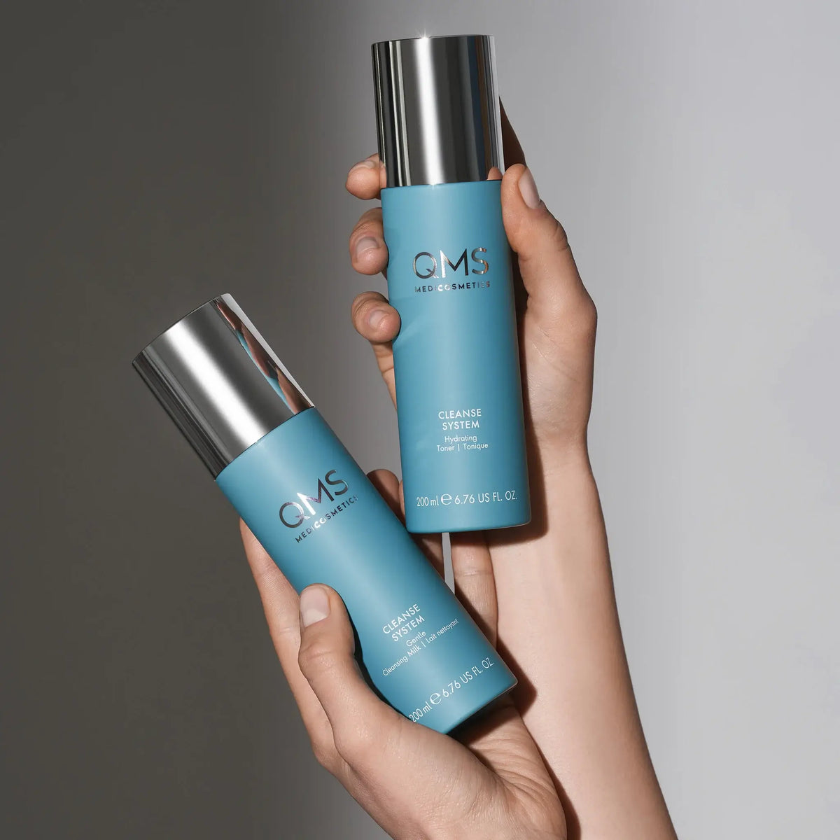 Zwei Hände halten blaue Flaschen von QMS Med Cosmetics Cleanse System und Cleanser, beide mit silbernen Kappen, vor einem neutralen Hintergrund.