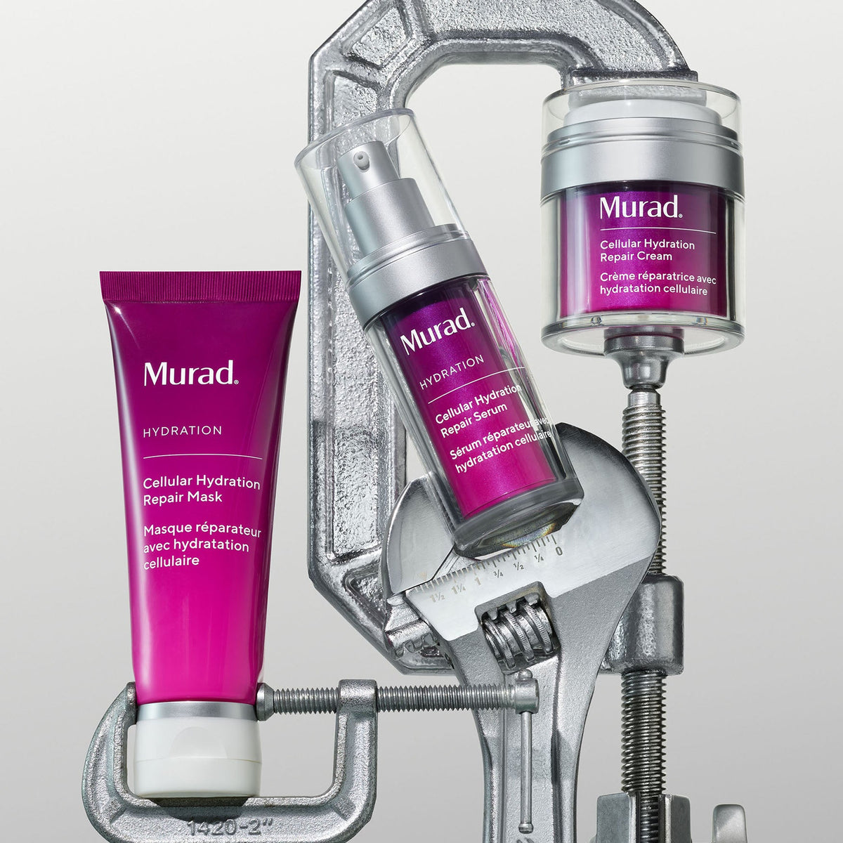Drei Murad Hydration Hautpflegeprodukte - eine Reparaturmaske, ein Serum und eine Creme - werden von einer großen Metallklammer und einem Schraubenschlüssel vor einem schlichten, hellen Hintergrund gehalten.