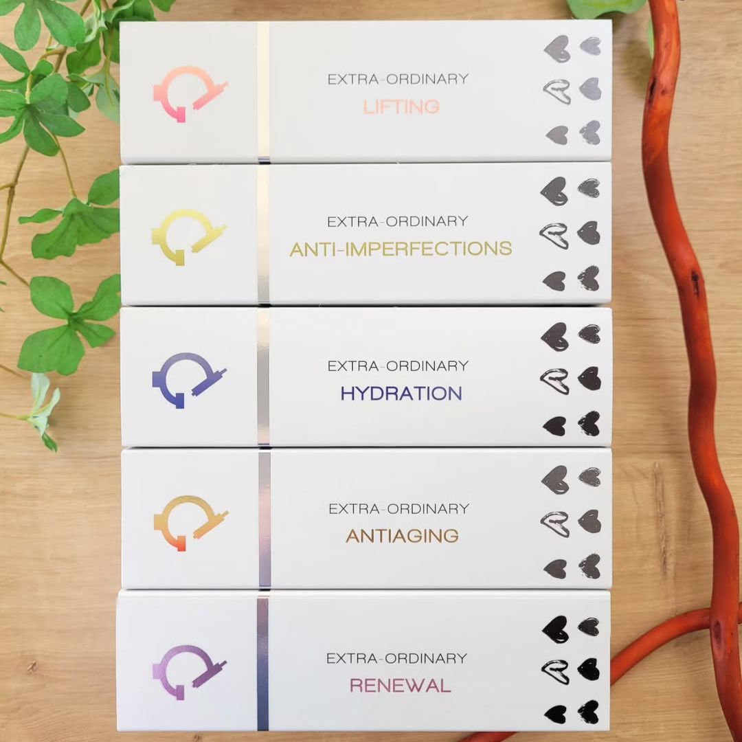 Fünf gestapelte Hautpflegeproduktboxen mit den Aufschriften Lifting, Anti-Imperfections, Hydration, Antiaging und Renewal auf einer hölzernen Fläche mit grünen und orangefarbenen Pflanzenstängeln daneben.