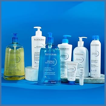 Eine Kollektion von Bioderma Atoderm Hautpflegeprodukten, einschließlich Flaschen mit Pumpen und Tuben, wird auf einem blauen Hintergrund mit weißen Marmorplattformen präsentiert.
