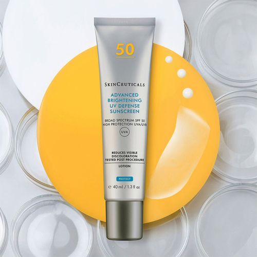 Eine Tube SkinCeuticals Advanced Brightening UV Defense Sunscreen SPF 50 liegt auf einem gelben und weißen Hintergrund, daneben sind Sonnenschutzlotion und -tropfen abgebildet.