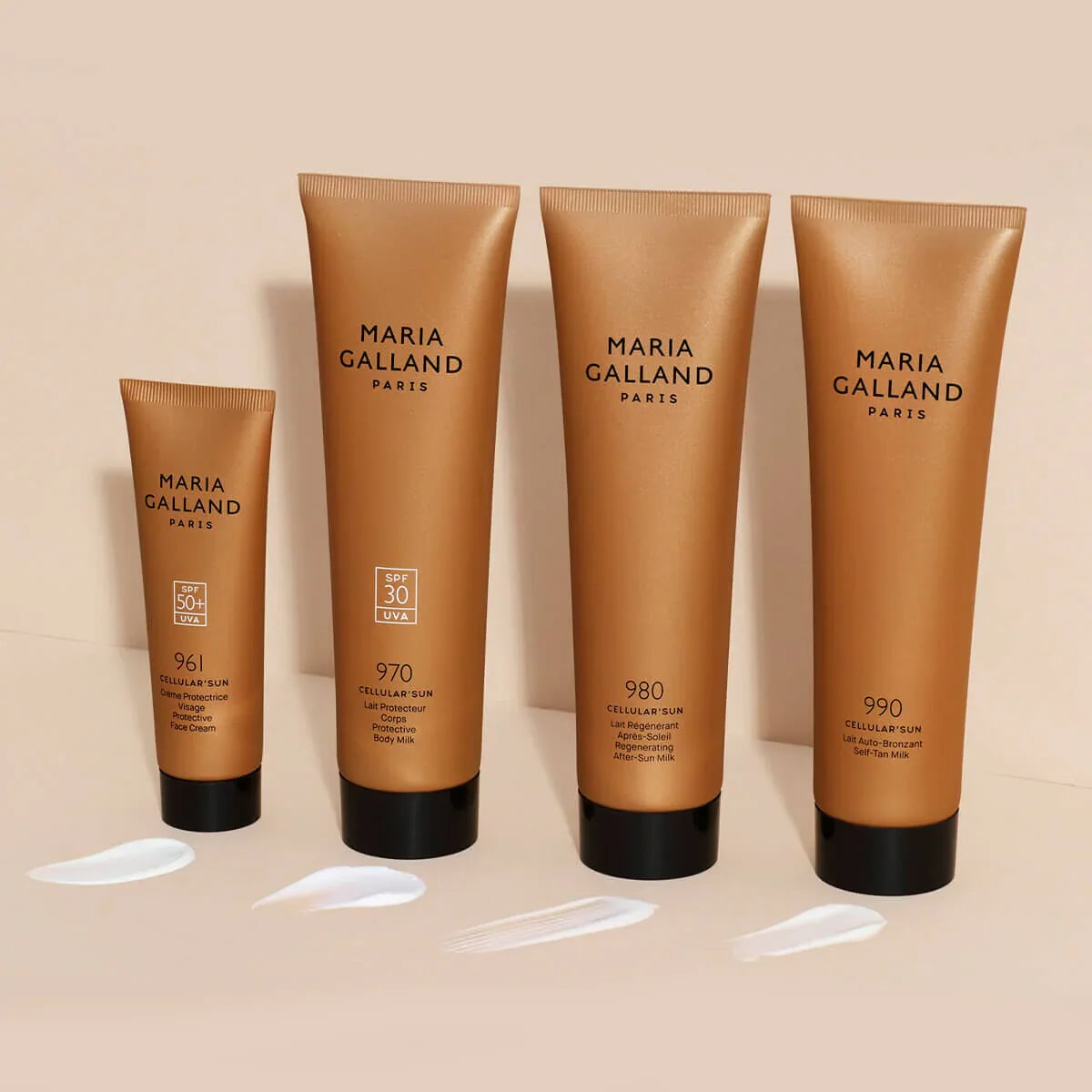 Vier goldene Tuben mit Hautpflegeprodukten von Maria Galland Paris stehen aufrecht auf einer beigen Fläche, vor denen sich jeweils ein Stück weiße Creme befindet. Die Tuben sind unterschiedlich groß.