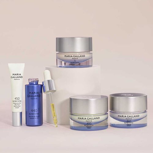 Fünf Hautpflegeprodukte von Maria Galland Paris, darunter Cremes in runden Tiegeln, ein Serum in einer blauen Tropfflasche und eine Augencreme in einer Tube, sind auf einem hellen Hintergrund mit einem weißen Block angeordnet.