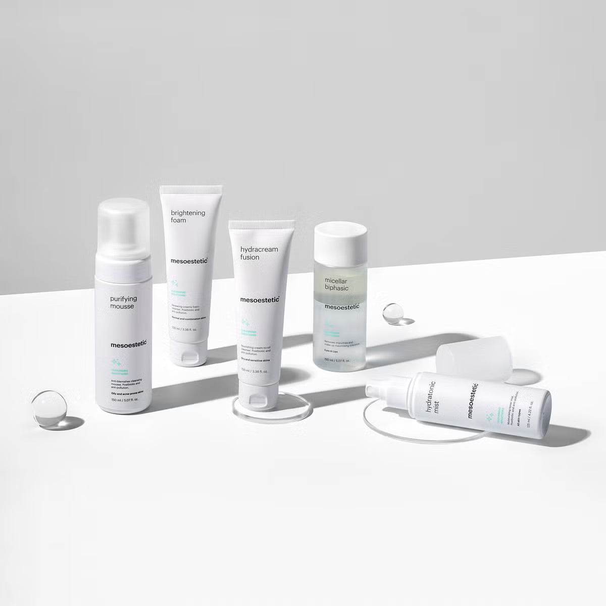 Fünf weiße Flaschen und Tuben von Hautpflegeprodukten mit der Aufschrift mesoestetic sind auf einer weißen Fläche mit klaren Tropfen und subtilen Schatten vor einem hellgrauen Hintergrund angeordnet.