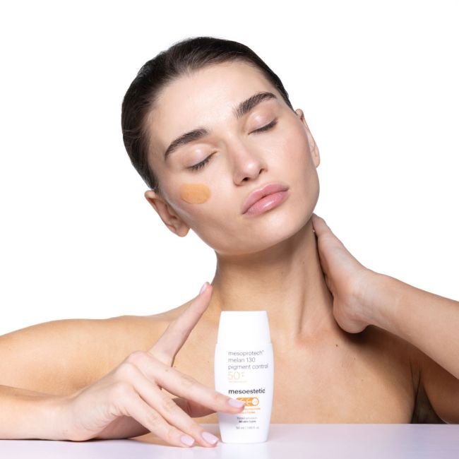 Eine Frau mit geschlossenen Augen trägt getönte Sonnencreme auf ihre Wange auf und hält eine Flasche mit der Aufschrift mesoestetic mesoprotech melan 130 pigment control SPF 50+ vor einem weißen Hintergrund.