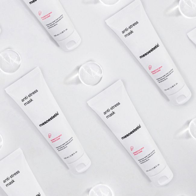 Mehrere weiße Röhrchen mit der Aufschrift mesoestetische Anti-Stress-Maske sind in einem diagonalen Muster auf einem hellen Hintergrund mit verstreuten klaren runden Objekten angeordnet.