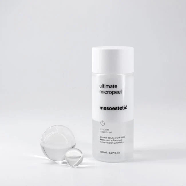 Ein weißes Fläschchen mit Mesoestetic Ultimate Micropeel steht aufrecht auf einer weißen Fläche, daneben zwei klare Glaskugeln, alles vor einem schlichten weißen Hintergrund.