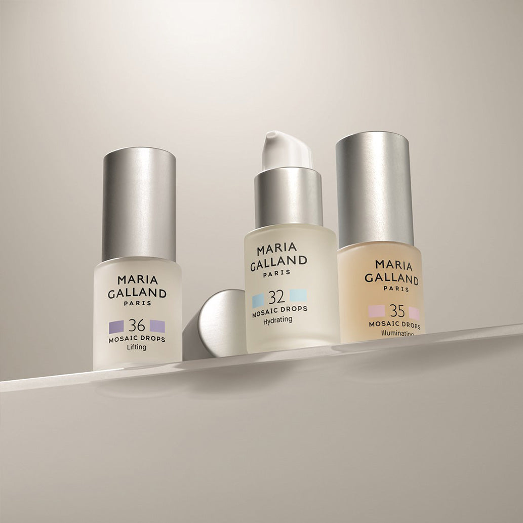 Drei Flaschen Maria Galland Paris Mosaic Drops (36 Lifting, 32 Hydrating mit Pumpe und 35 Illumination) sind auf einem weißen Regal vor einem neutralen Hintergrund ausgestellt.