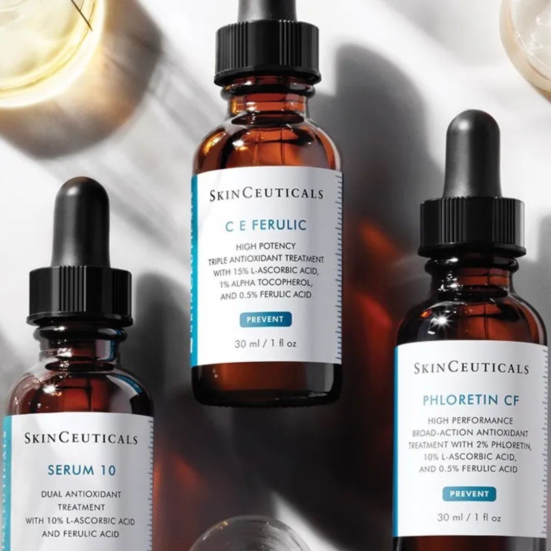 Drei bernsteinfarbene Tropfflaschen mit SkinCeuticals-Seren (CE Ferulic, Phloretin CF und Serum 10) sind auf einer weißen Fläche mit Schatten und Teilansichten der anderen Produkte angeordnet.