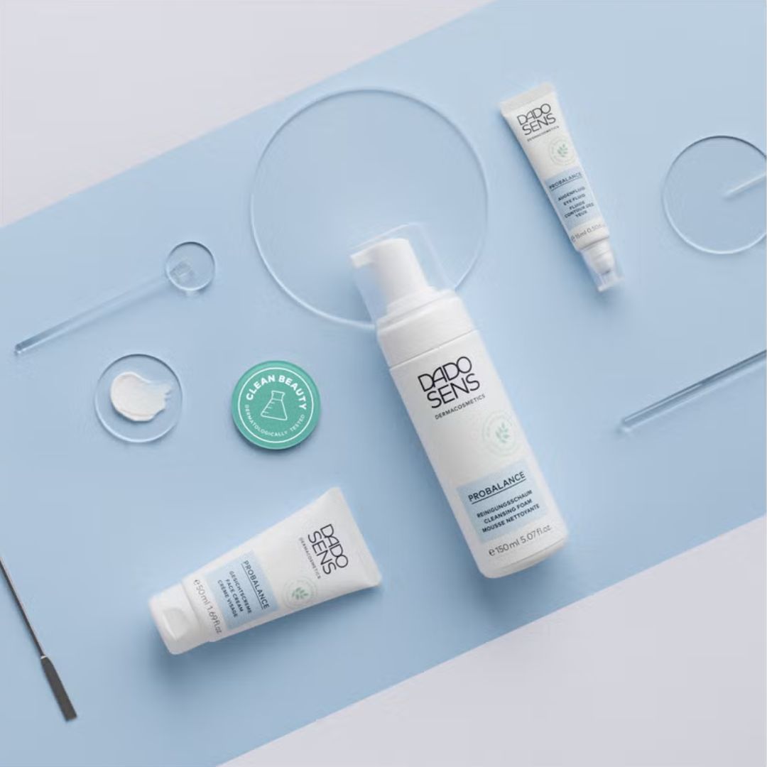 Die Hautpflegeprodukte von Dado Sens Probalance, darunter ein Reinigungsschaum, eine Creme und eine Tube, sind auf einer hellblauen Fläche mit Glaskreisen, Applikatoren und einer Clean Beauty-Plakette angeordnet.