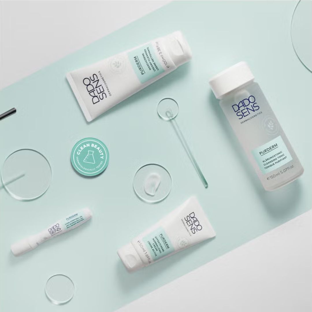 Eine flache Lage von Dado Sens Purderm Hautpflegeprodukten auf einer hellblauen Oberfläche mit klaren runden Glasstücken und einem Clean Beauty Abzeichen. Die Produkte umfassen Cremes, eine Flasche und eine Tube.
