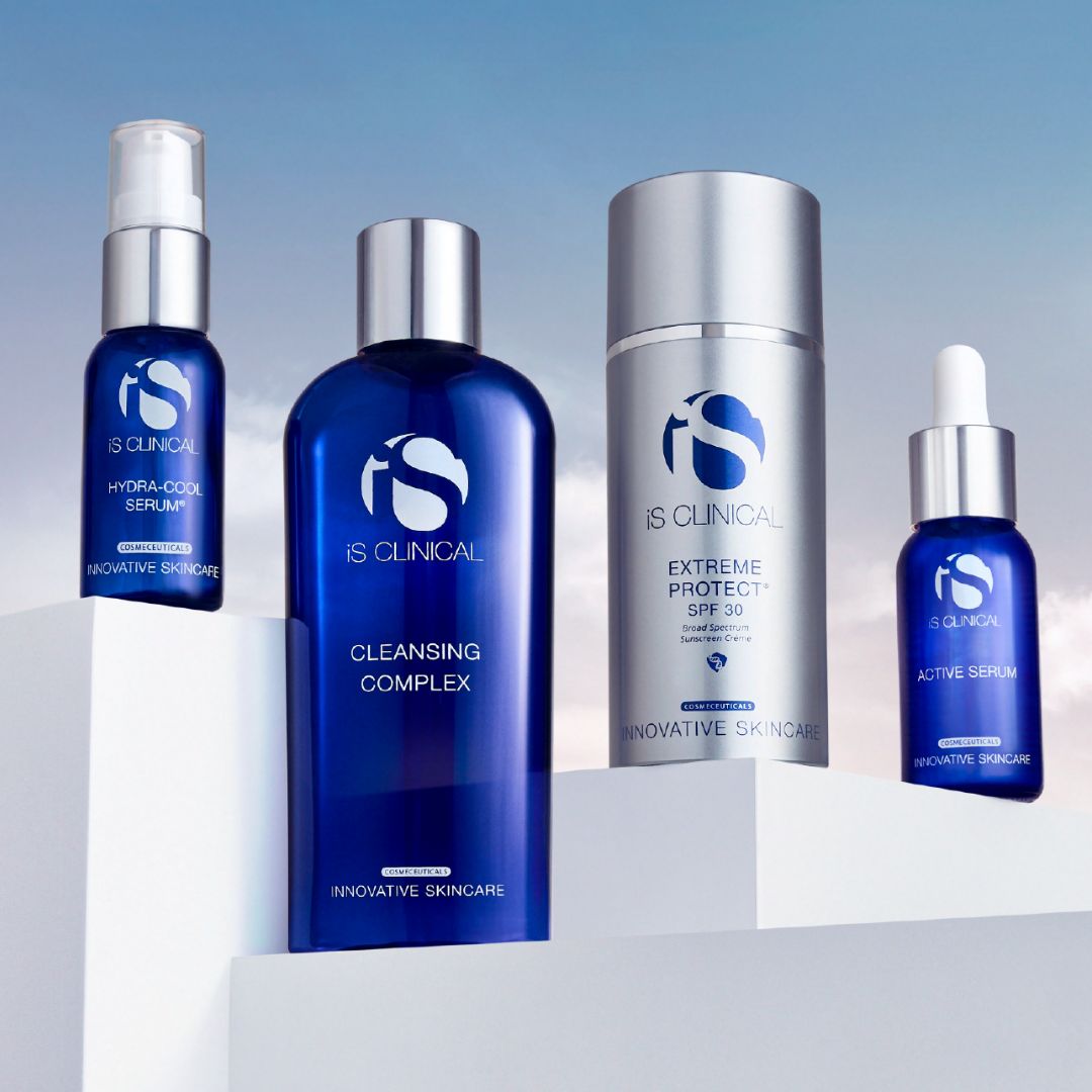 Vier blaue Hautpflegeflaschen und -behälter mit der Aufschrift iS Clinical sind auf weißen Blöcken vor einem himmelblauen Hintergrund abgebildet. Zu den Produkten gehören Seren, ein Reinigungskomplex und eine SPF-Lotion.