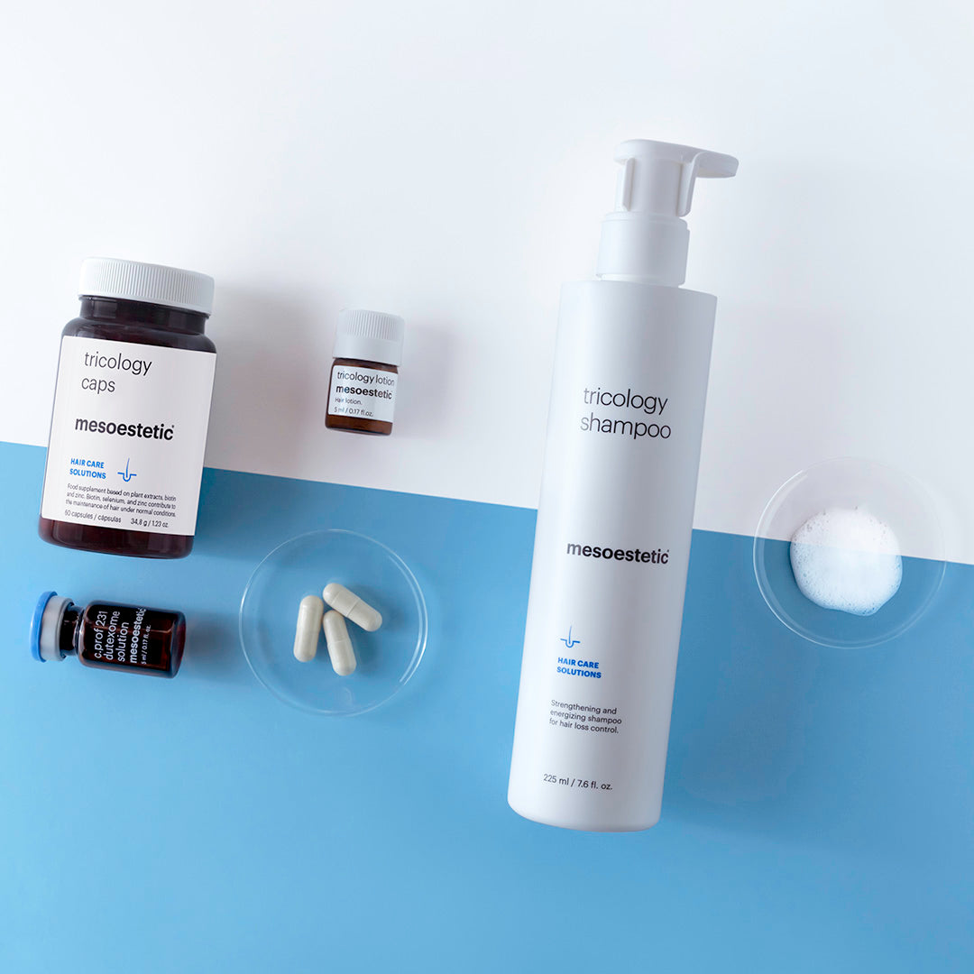 Eine flache Lage von Mesoestetic-Haarpflegeprodukten umfasst eine Shampooflasche, Kapseln für Nahrungsergänzungsmittel, zwei kleine Medizinflaschen und zwei Glasschalen mit Kapseln und Pulver, die auf einem blauen und weißen Hintergrund dargestellt sind.