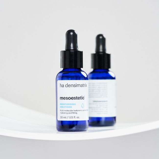 Zwei blaue Tropfflaschen mit mesoestetic ha densimatrix Serum stehen auf einer weißen Fläche, wobei eine Flasche mit dem Produktetikett nach vorne zeigt und die andere zur Seite gedreht ist.