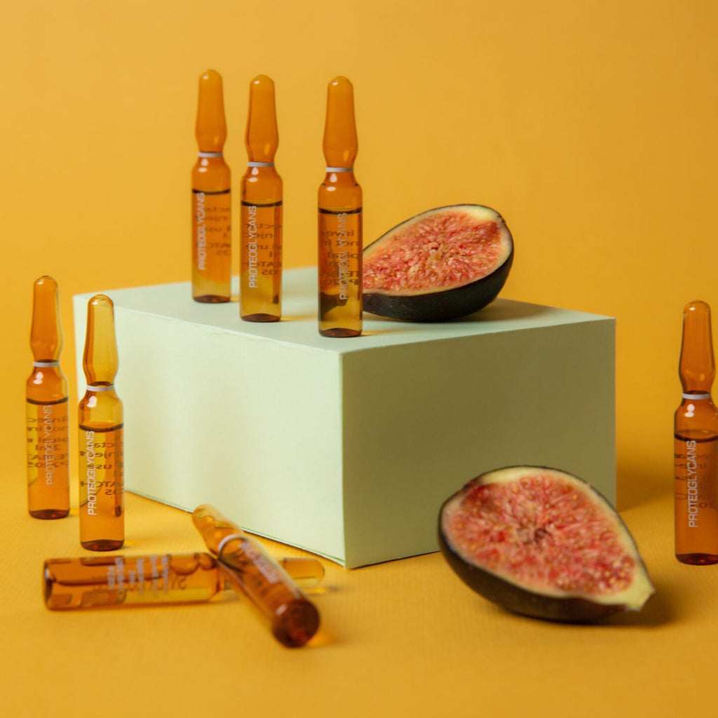 Sechs Alchemy - Proteoglycans Plus Ampullen von Alchemy Care Cosmetics und zwei halbierte Feigen sind auf und um einen hellgrünen rechteckigen Block angeordnet, der auf einem einfarbigen gelben Hintergrund steht.