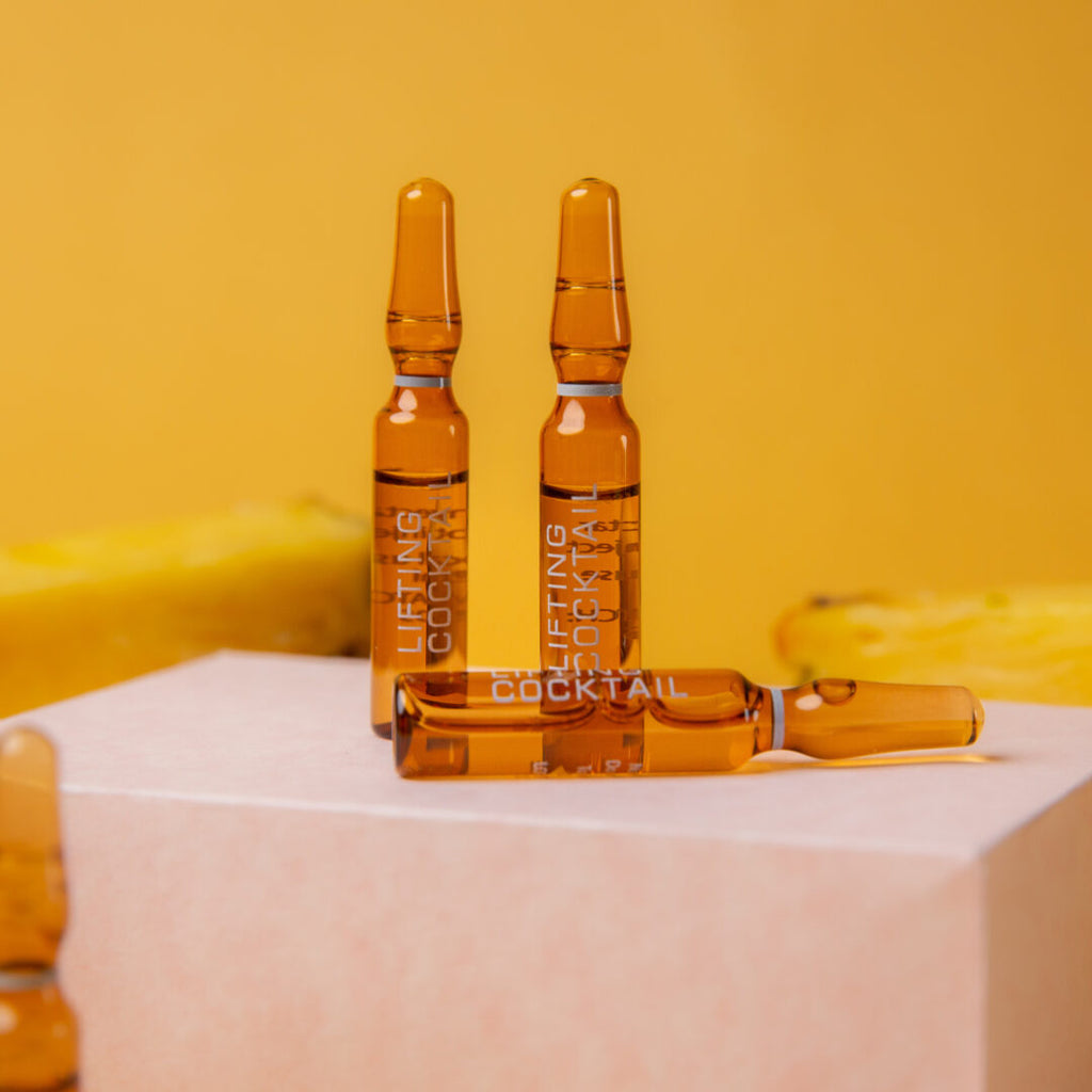 Drei Braunglasampullen mit der Aufschrift Alchemy - Anti Aging Lifting Cocktail von Alchemy Care Cosmetics sind auf einem gelben Hintergrund abgebildet, wobei zwei aufrecht stehen und eine flach auf einer rosafarbenen rechteckigen Fläche liegt.
