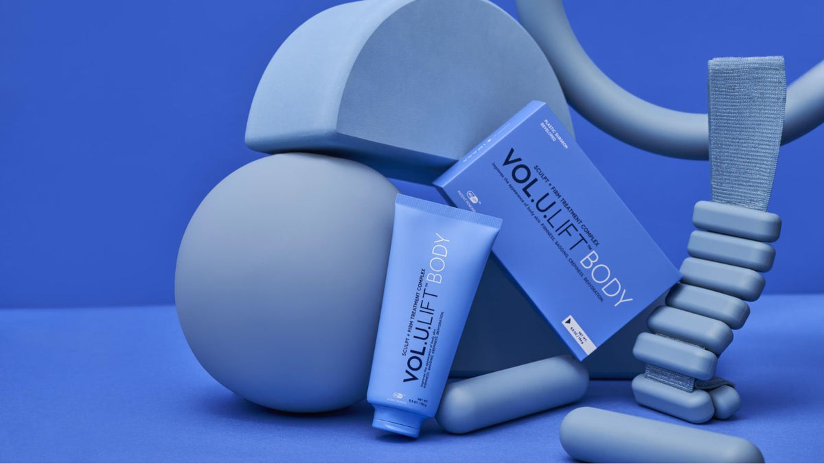 Eine Tube und eine Schachtel Volulift Body Sculpt + Firm Treatment Complex vor einem blauen Hintergrund mit abstrakten Formen.